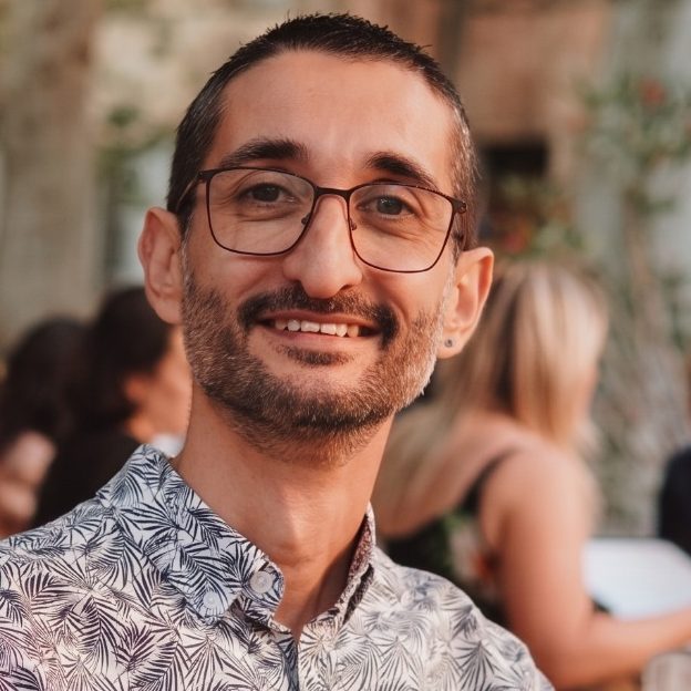 PARTENAIRE Polymathia Group Joël Escrich souriant avec des lunettes, portant une chemise à motifs, en arrière-plan flou.