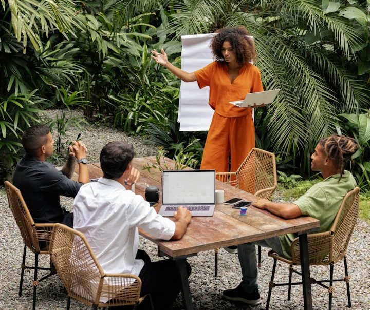 Une femme présente devant trois personnes lors d'une réunion en extérieur, entourée de verdure.