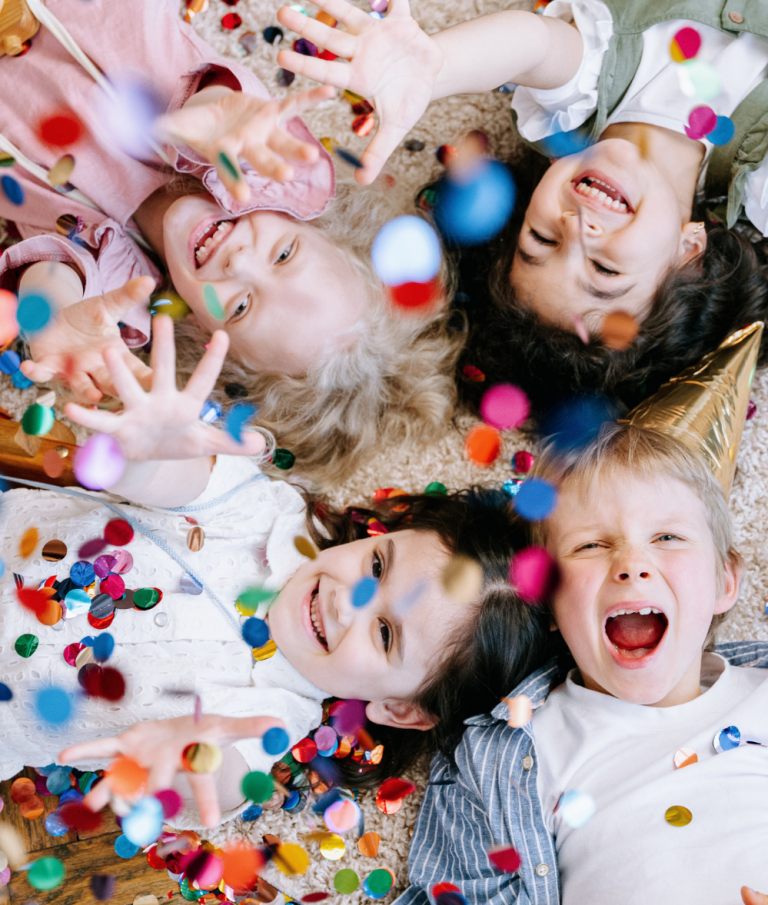Quatre enfants joyeux entourés de confettis colorés, souriant à la caméra.