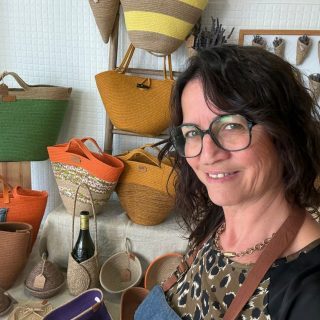 PARTENAIRE Polymathia Group Femme souriante devant des paniers en osier colorés et divers objets artisanaux.