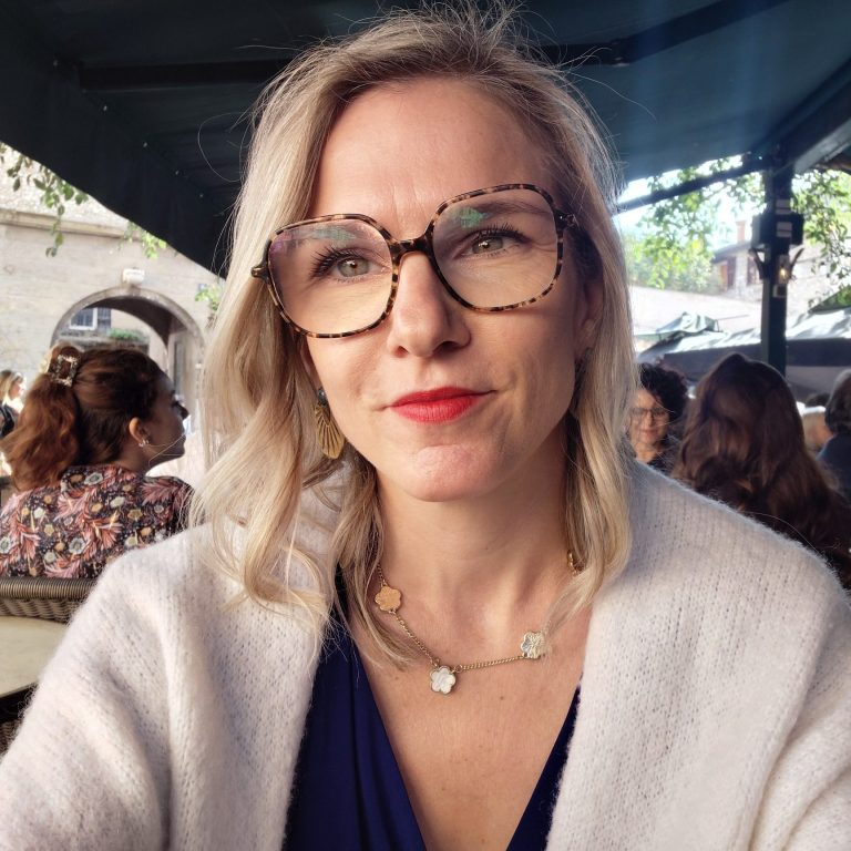 Femme souriante portant des lunettes, vêtue d'un haut noir, avec un fond artistique.