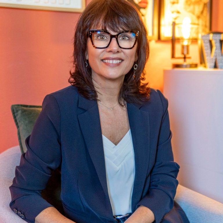 Nikolina Popovski Coach en vente Femme souriante portant des lunettes, vêtue d'un haut noir, avec un fond artistique.