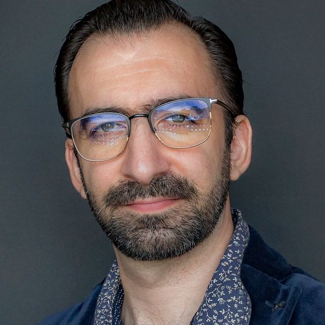 PARTENAIRE Polymathia Group Homme en costume gris avec une barbe, souriant devant un fond gris.
