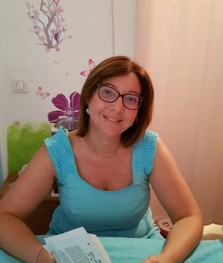 Femme souriante portant un haut bleu, assise dans une chambre décorée.