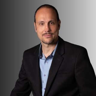 PARTENAIRE Polymathia Group Homme en costume noir avec une chemise bleue, souriant, fond gris dégradé.