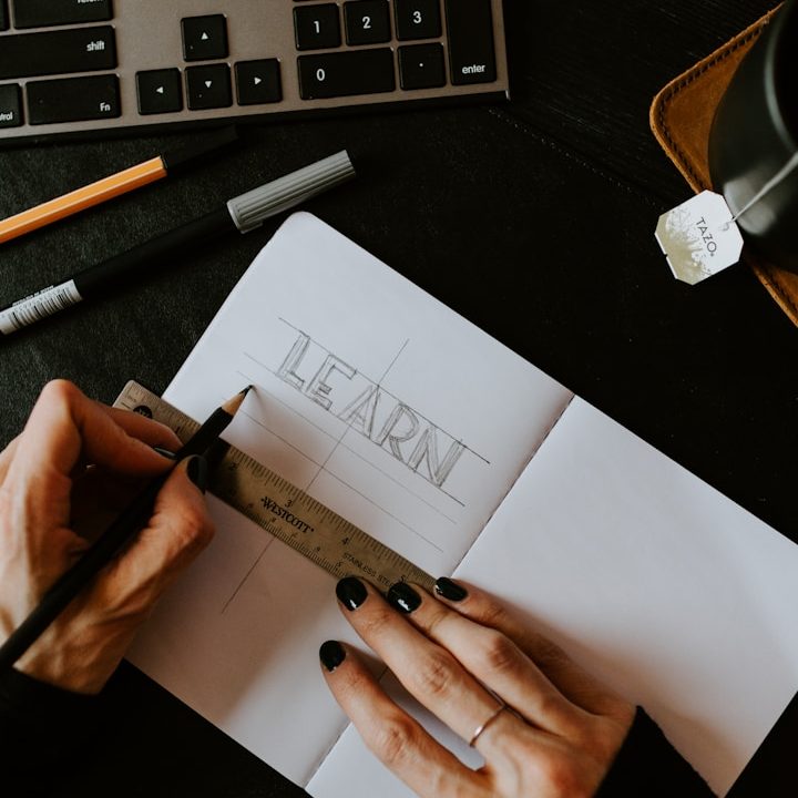 Main en train de dessiner "LEARN" dans un carnet avec une règle et des stylos.