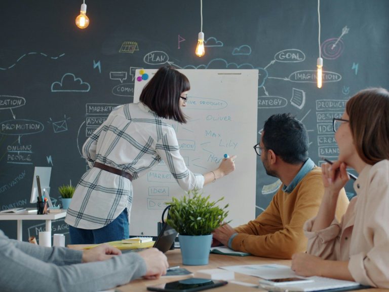 La Polymathia Group Academy Une personne présente des idées sur un tableau, pendant que d'autres écoutent attentivement.